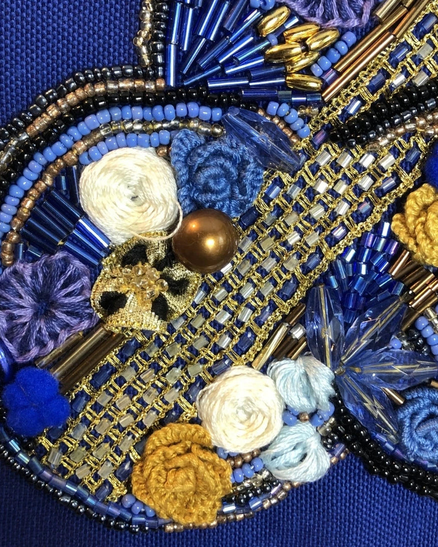 Close-up of an ornate beaded and embroidered decorative motif on a blue fabric background, featuring blue and gold beads, white and light blue rosettes, yellow flowers, a small skull detail, and a metallic gold center bead. A stylized 'YD' mark appears below the artwork.