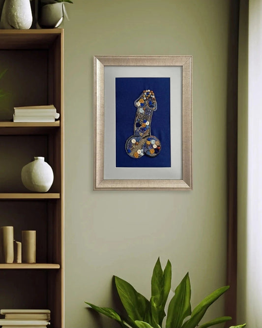 Framed wall art featuring a stylized blue body part with gold and silver spots, displayed on a sage green wall in a styled room setting with a wooden shelf and plants.