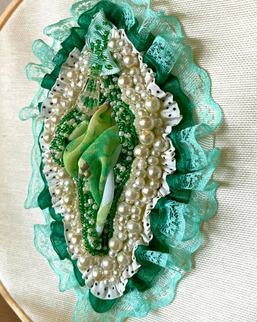 An ornate embroidered brooch featuring a green and white bird motif surrounded by white faux pearls, green beads, and layered green lace on a beige fabric background.