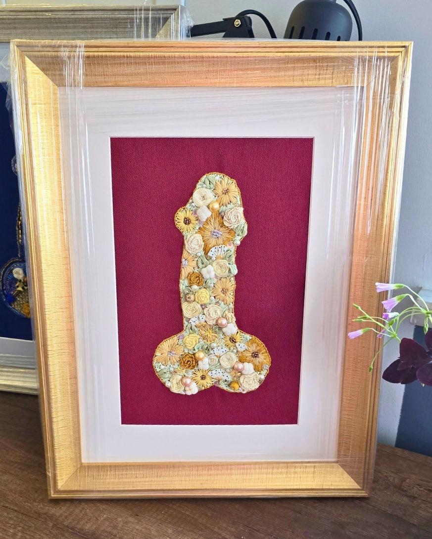 Framed art print featuring a stylized penis shape crafted from small yellow and white flowers on a deep red background, housed in a gold frame and displayed on a wooden surface.