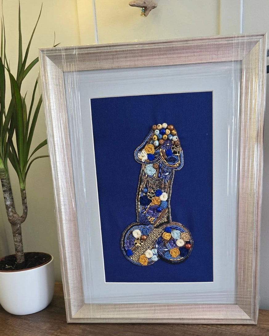 Framed art print featuring a stylized penis design made from blue, gold, and white beads, mounted on a dark blue background inside a light wood frame, displayed next to a potted plant.