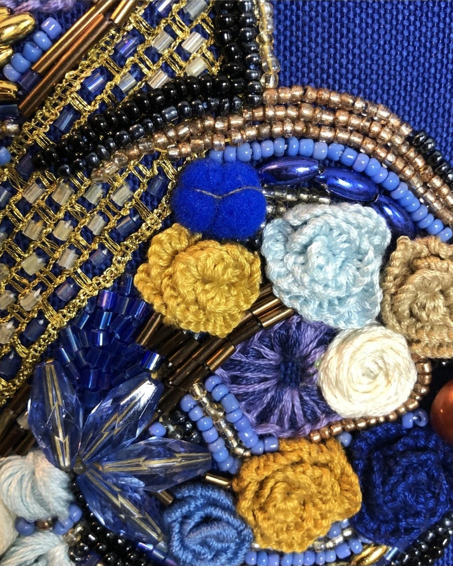 Close-up of an ornate beaded and embroidered accessory featuring blue beads, gold metallic details, crocheted fabric flowers in blue, yellow, white, and beige, and a glossy brown bead on a textured blue background.