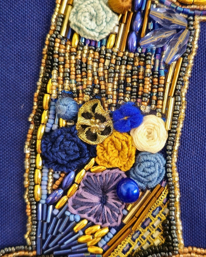 Close-up of a beaded garment or textile detail featuring blue, gold, purple, yellow, and cream floral and geometric beadwork on a dark blue fabric background.