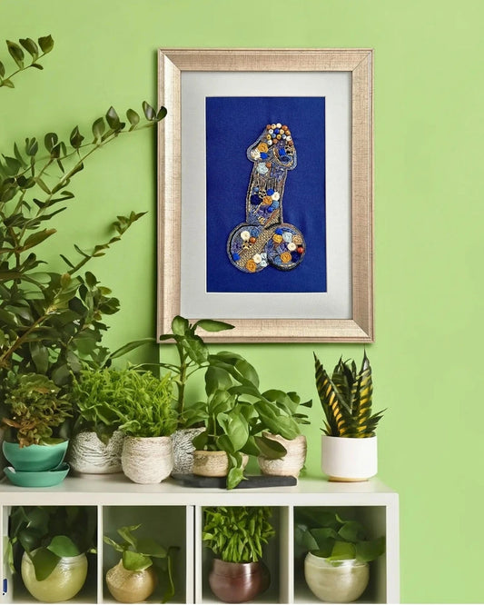 A framed art piece featuring a stylized blue and gold body illustration, displayed on a white shelf surrounded by potted green plants against a light green wall.