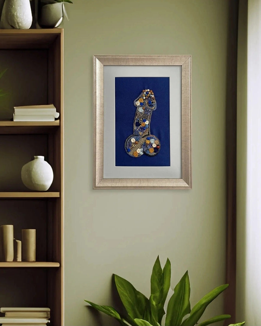 Framed wall art featuring a stylized blue body part with gold and silver spots, displayed on a sage green wall in a styled room setting with a wooden shelf and plants.