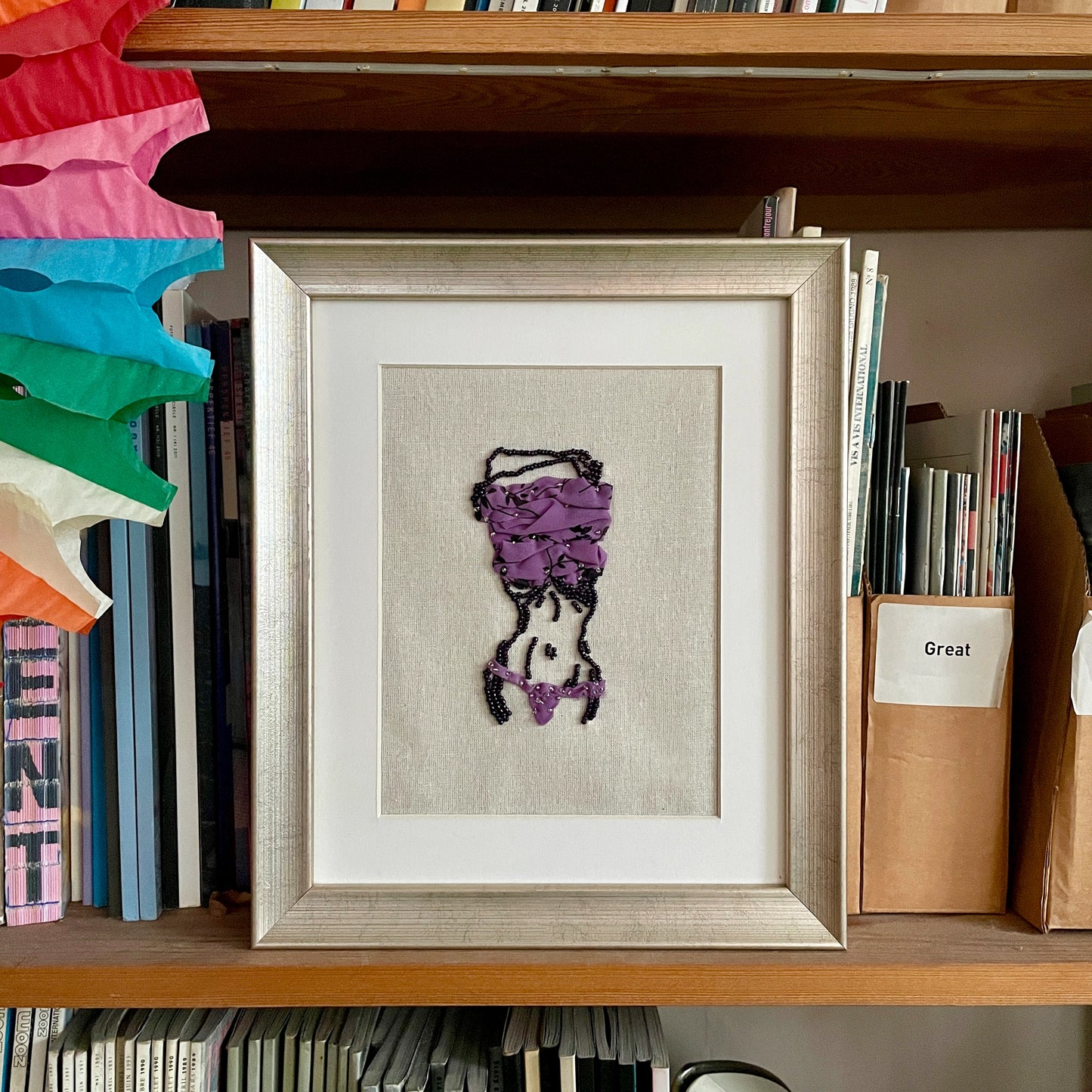 A framed piece of textile art featuring a stylized purple and black bird figure on a beige linen background, displayed on a wooden bookshelf next to books and decorative paper fans.